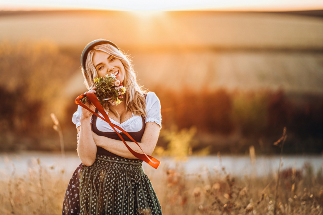 Lächelnde junge Frau im Dirndl mit Blumenstrauß auf einem Feld im Abendlicht – bayerisches Lebensgefühl zum Geburtstag feiern.